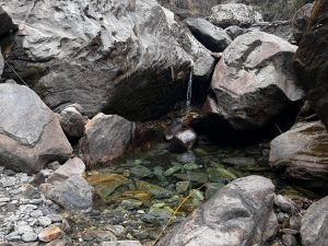 A rocky landscape featuring large boulders of various shapes and sizes arranged around a clear, shallow pool of water. 