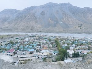 A panoramic view of a small town nestled between high mountains and a river. The town features a mix of buildings, including houses with colorful roofs and some larger structures.